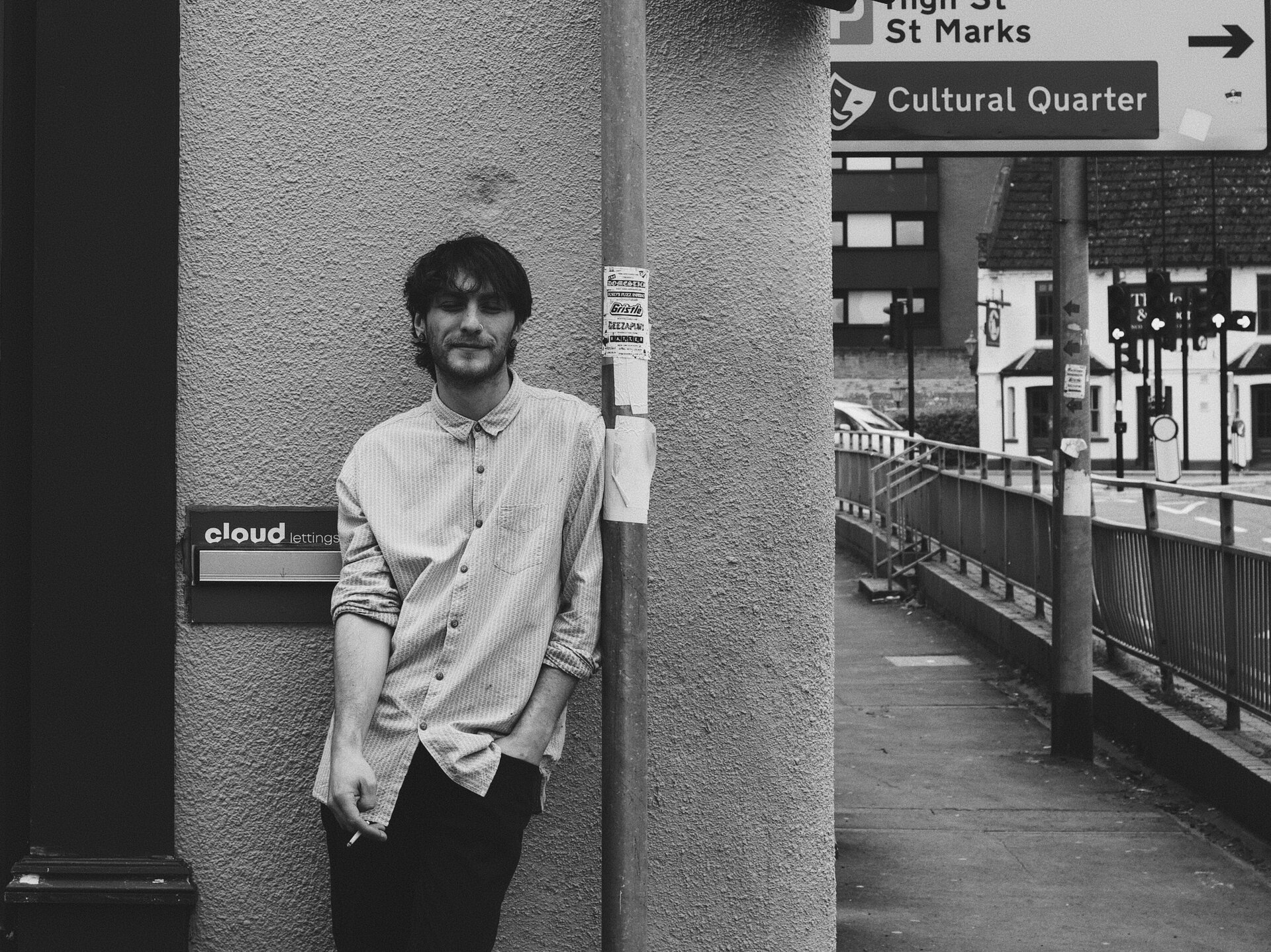 A man in black and white leaning against a pillar beside a street sign