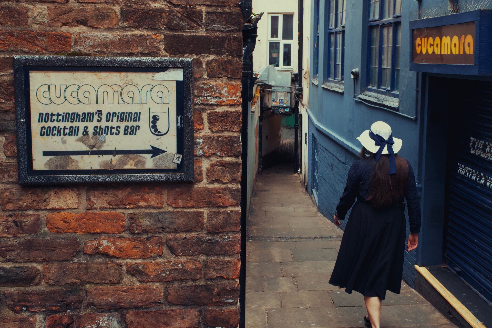 A woman in a white hat walking down a narrow alley past a cocktail bar sign